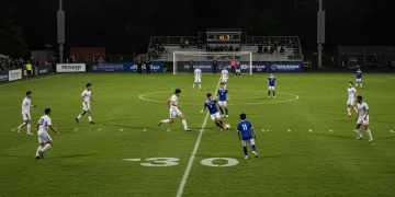 College soccer team executing a modern tactical formation on the field during a competitive match under stadium lights.